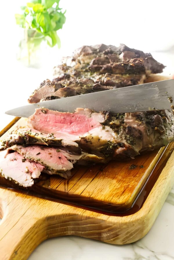 Knife slicing butterflied leg of lamb on wooden board.