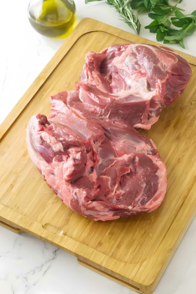 A raw leg of lamb butterflied on a cutting board with olive oil and herbs.