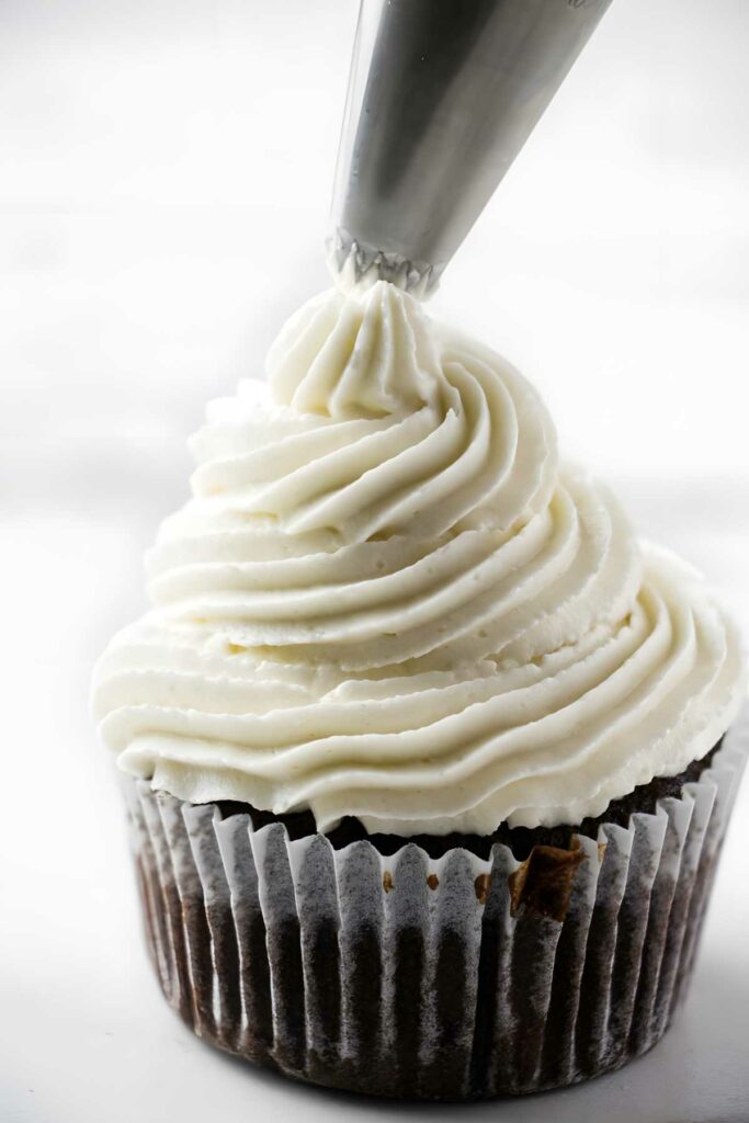 Chocolate cupcake being topped with white frosting from piping bag, white background.