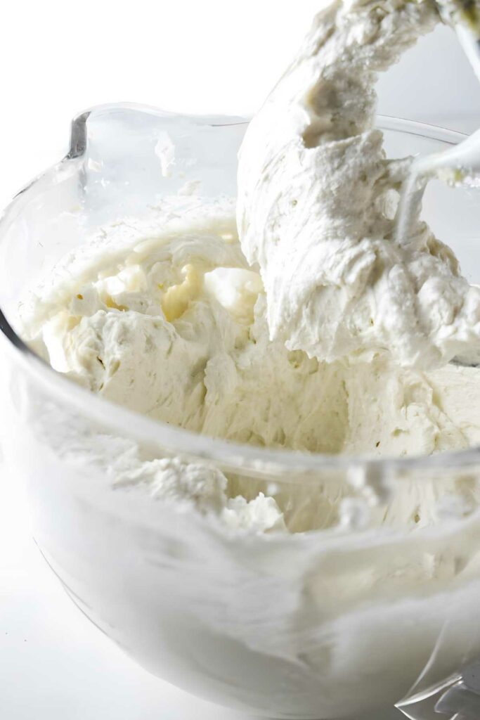 Close-up of stand mixer blending creamy white frosting in glass bowl.