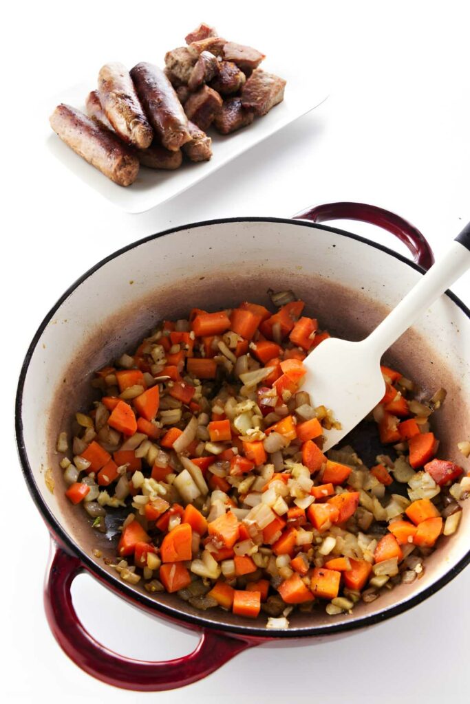 Red Dutch oven with sautéed vegetables, stirred; plated browned sausages in background.