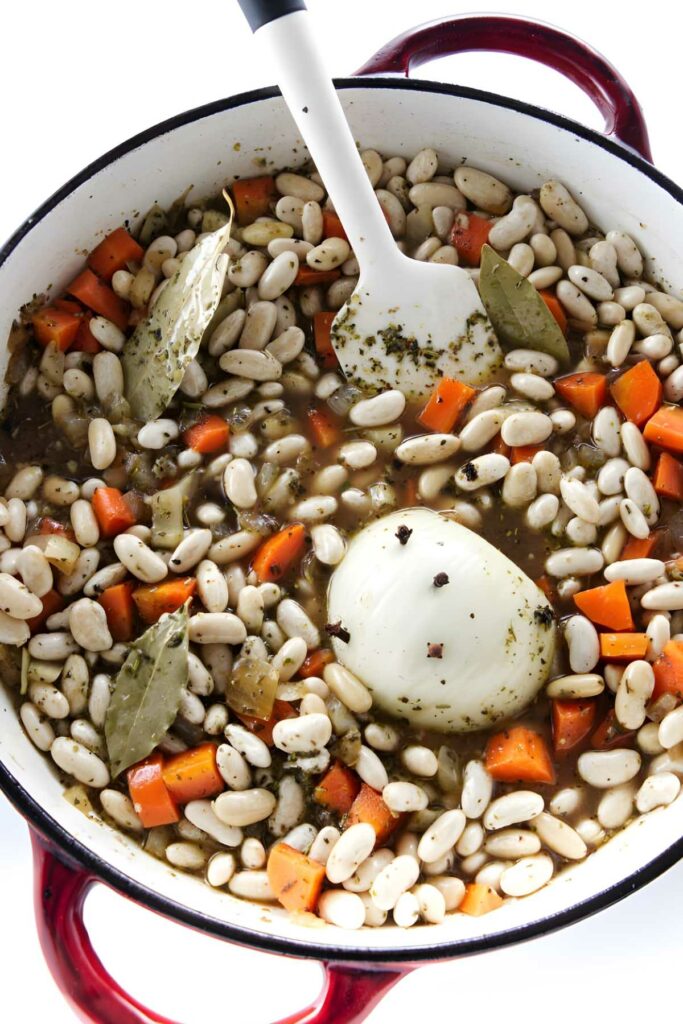 Red Dutch oven with beans, carrots, onion, and herbs in broth.
