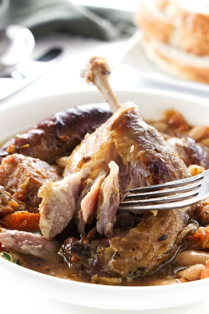 Fork pulls tender meat from duck leg in stew with beans and vegetables.