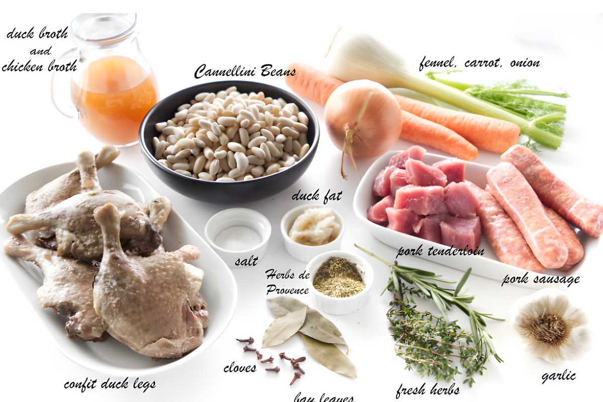 Cassoulet ingredients arranged on white surface, including meats, beans, vegetables, herbs.