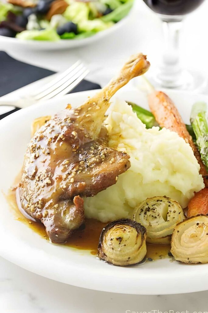 Roasted duck leg with sides on plate; wine and salad in background.