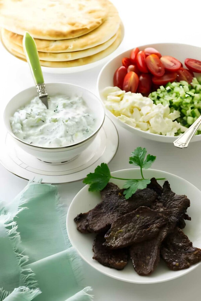 Grilled meat with parsley, tzatziki, vegetables, and flatbreads on white table.