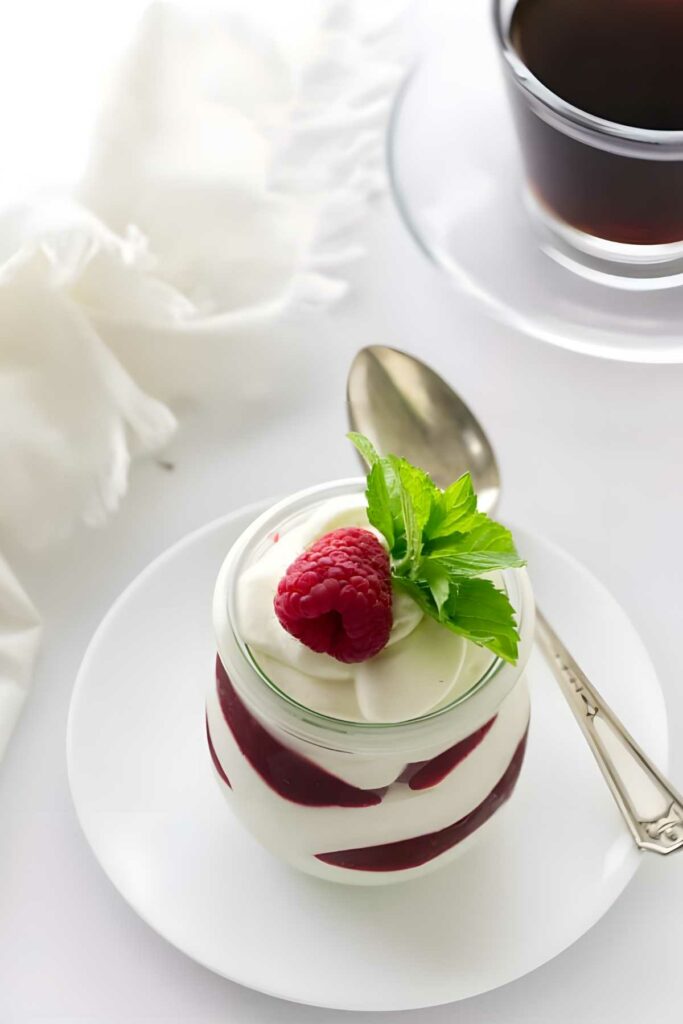 Layered dessert jar with raspberry, mint, coffee cup and spoon nearby.