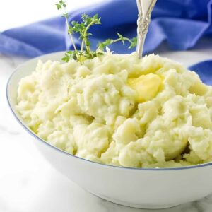 Bowl of mashed potatoes with butter, herbs, and silver spoon on marble.