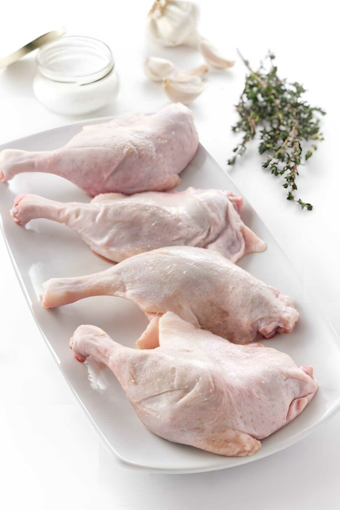 Three raw duck legs on plate with garlic, thyme.