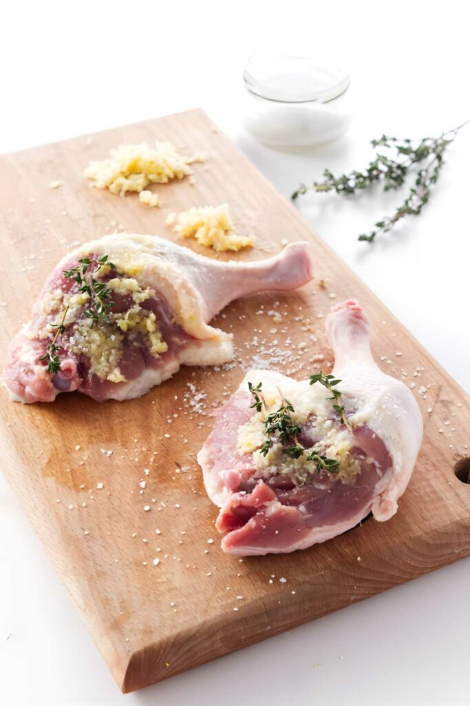 Two raw duck legs on a board with salt, garlic, and thyme.