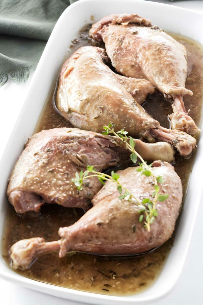 Four cooked duck legs with brown sauce, garnished with fresh thyme.