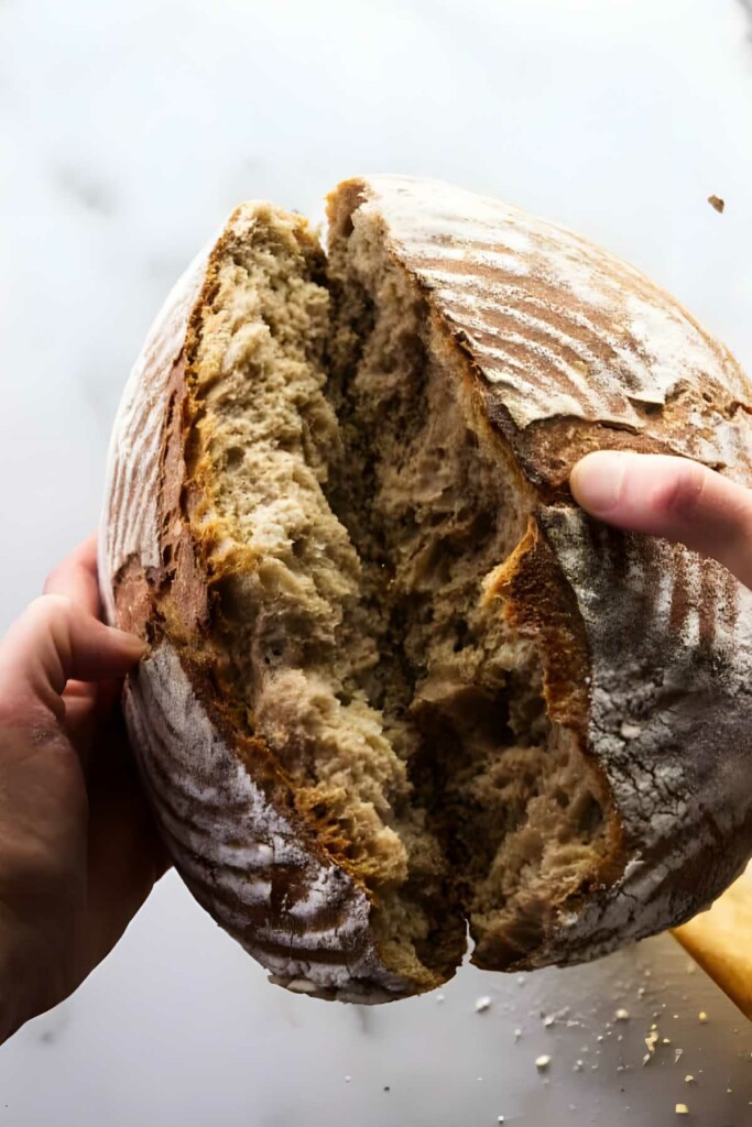 Hands breaking open rustic loaf, revealing airy crumb and patterned crust.
