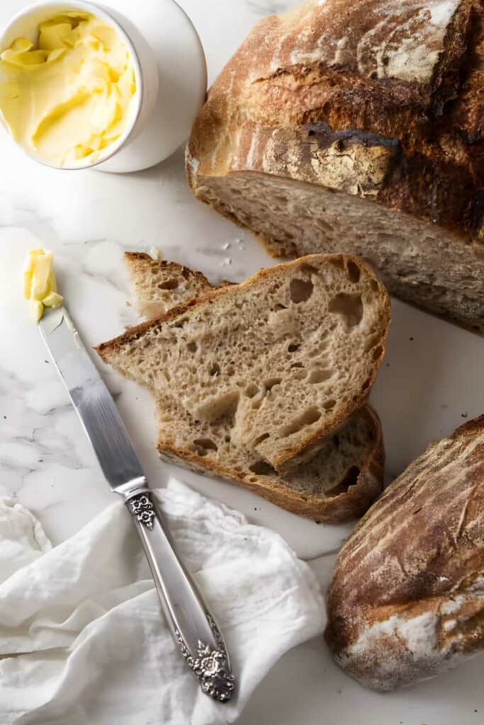 Sourdough loaf with sliced pieces, knife, butter bowl, on marble surface.