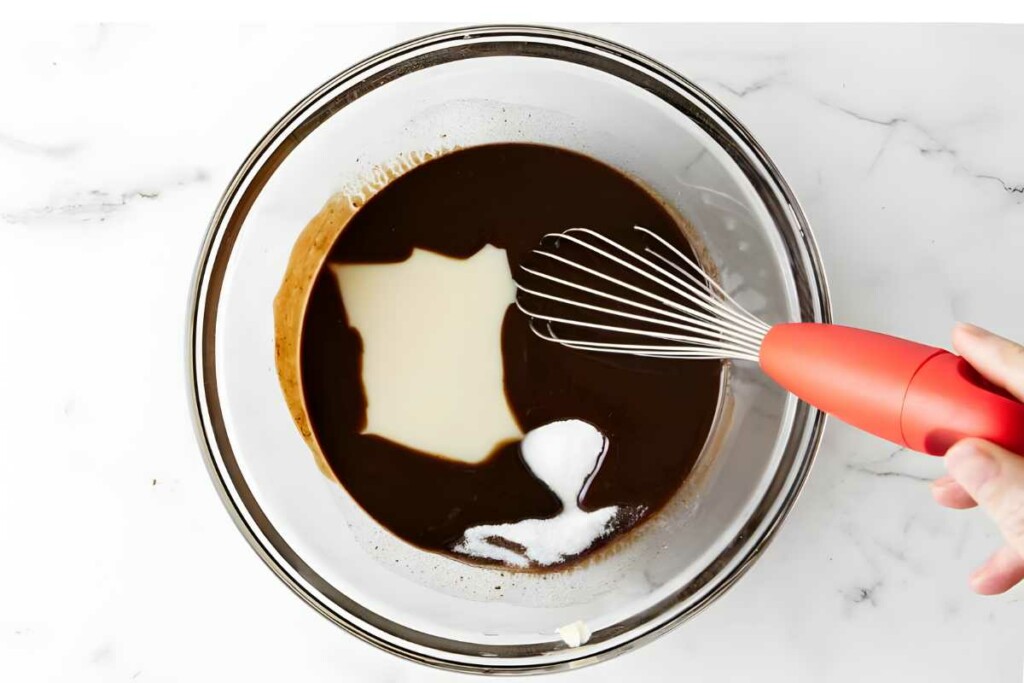 Hand holding whisk mixes liquids and sugar in glass bowl.