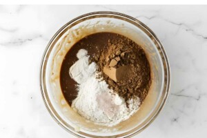 Glass bowl with brown batter, flour, and cocoa.