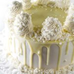 White cake with white frosting, rosettes, chocolate drip on ornate stand.
