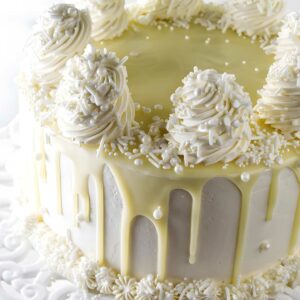 White cake with white frosting, rosettes, chocolate drip on ornate stand.