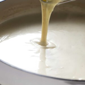 Whisk lifting creamy white sauce above a saucepan on light countertop.