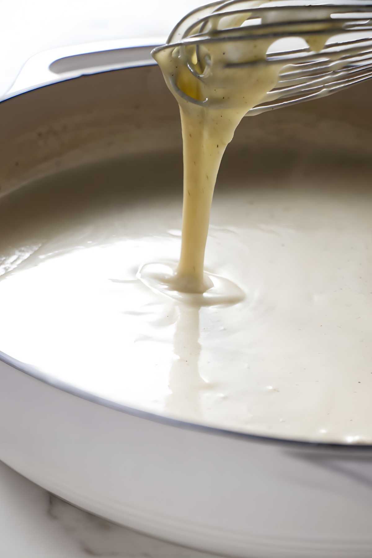 Whisk lifting creamy white sauce above a saucepan on light countertop.