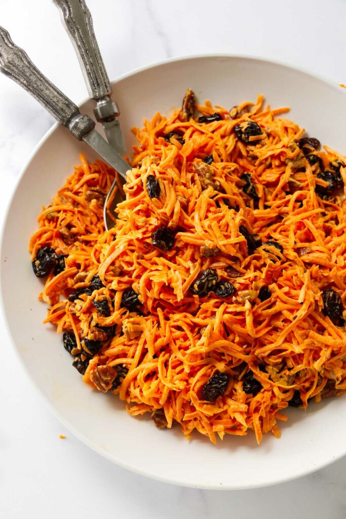 Carrot salad with raisins in a bowl with serving spoons.