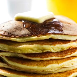 Stack of pancakes with butter and syrup, orange juice and egg behind.