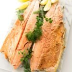A baked salmon with tail, garnished with herbs and lemon wedges.