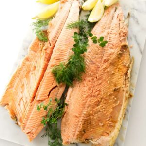Two cooked salmon fillets with tail, garnished with herbs and lemon wedges.