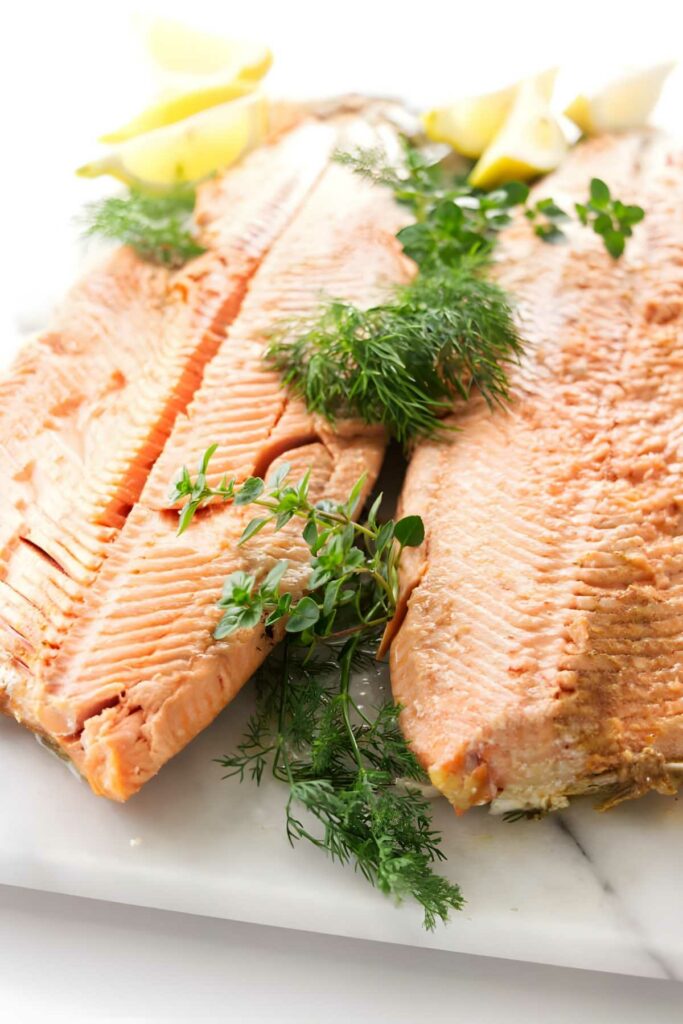 Two cooked salmon fillets with dill, thyme, and lemon wedges on marble.