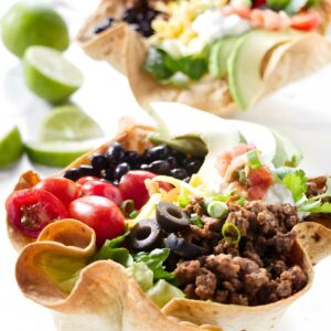 Two tortilla bowls filled with colorful taco salad, limes in background.