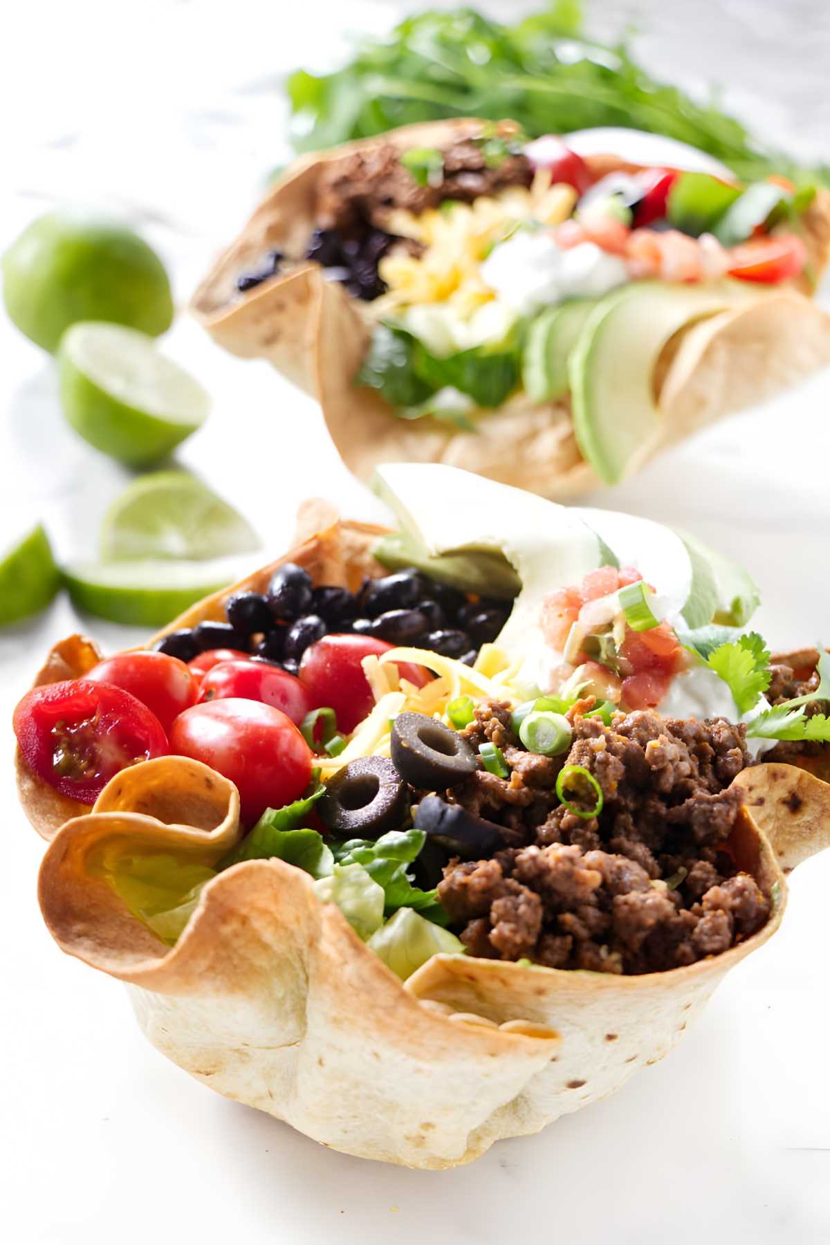 Two tortilla bowls filled with colorful taco salad, limes in background.