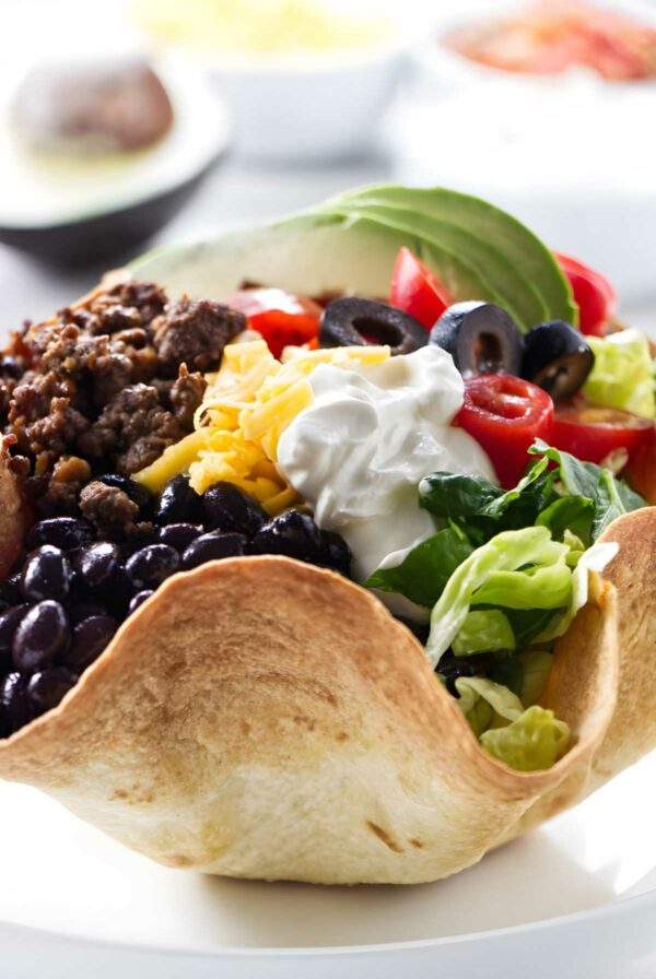 Taco salad in tortilla bowl with beef, beans, veggies, avocado, sour cream.