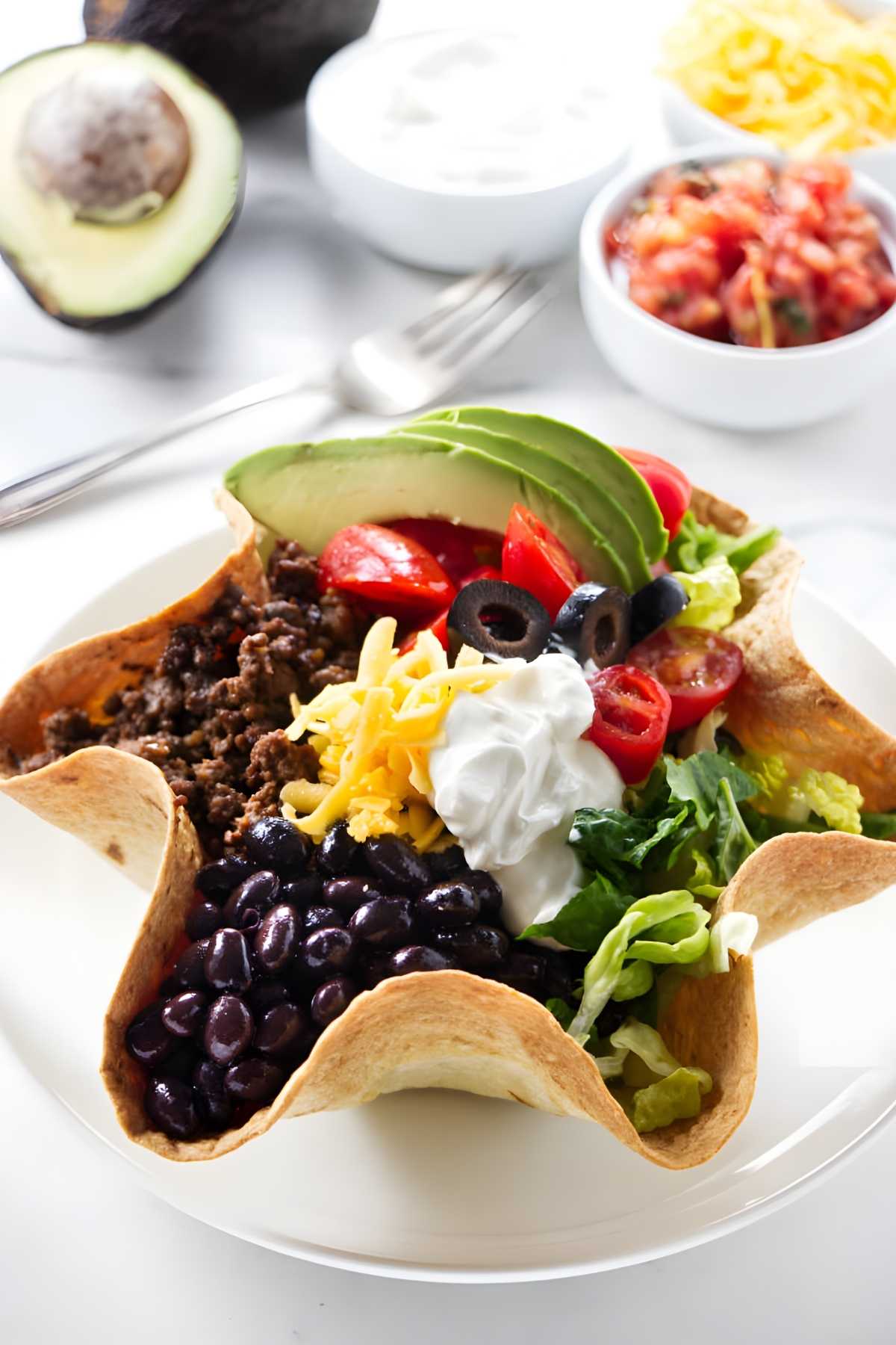 Taco salad in tortilla bowl with toppings; salsa and sides in background.
