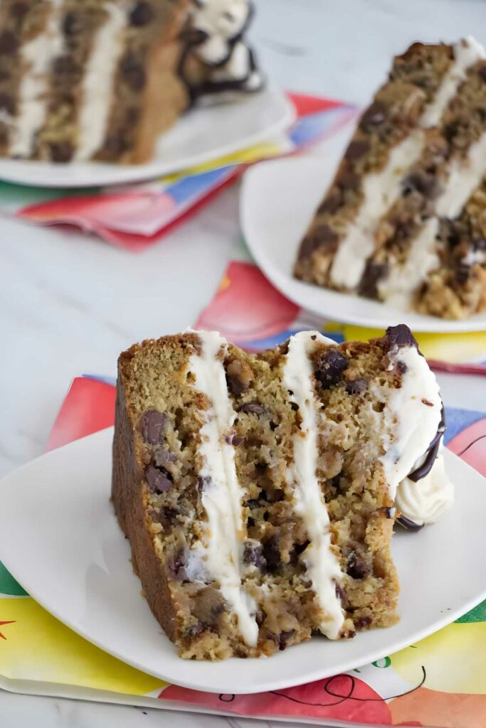 Slice of layered cookie cake with chocolate chips and white frosting on plate.