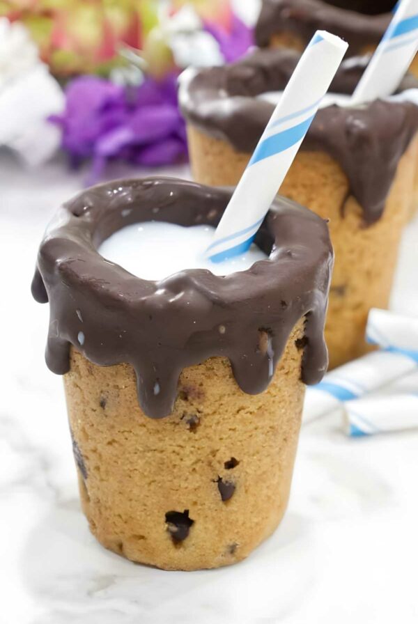 Cookie cup with chocolate, milk, straw; more cups and flowers behind.