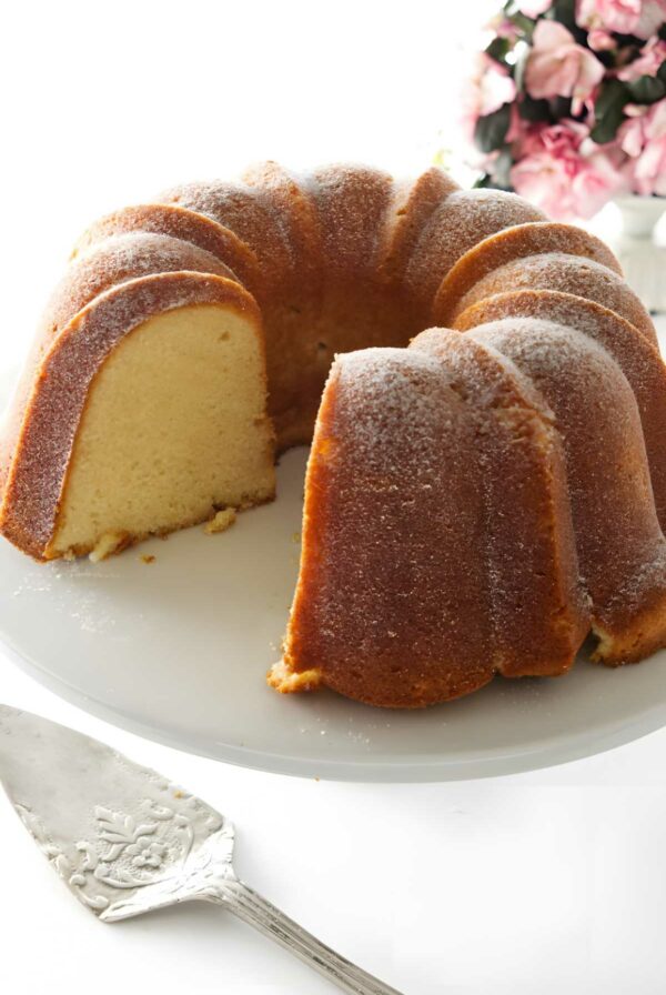 Golden-brown cold oven pound cake on stand, one slice removed, flowers behind.