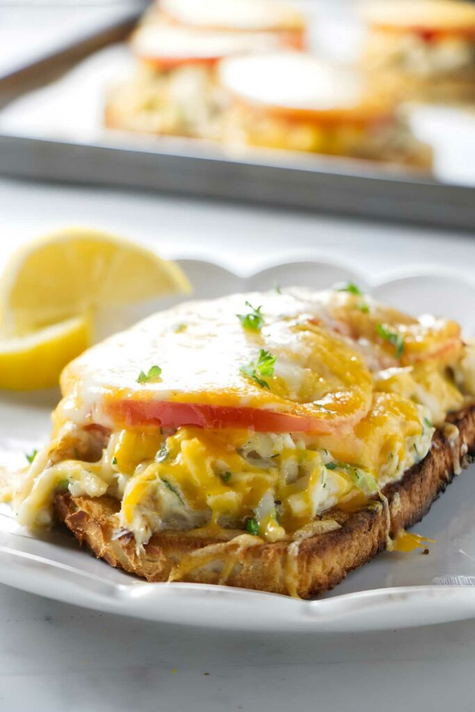 Cheesy crab toast with tomatoes, melted cheese, herbs, and lemon wedge.