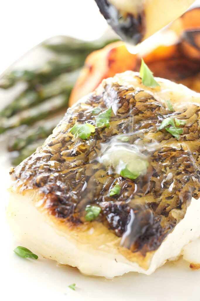 Close-up of crispy fish fillet with herbs, sauce, and blurred vegetables.
