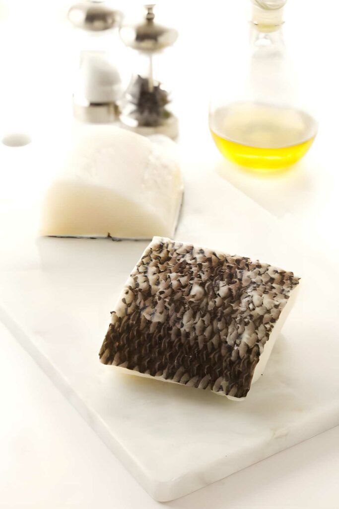 Raw fish fillet with dark skin on white cutting board, kitchen items behind.