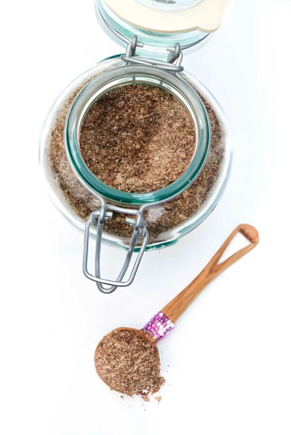 Open glass jar and wooden spoon with ground flaxseed on white surface.