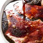Hand pours dark marinade over raw beef steaks.