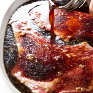 Hand pours dark marinade over raw beef steaks with garlic and spices.