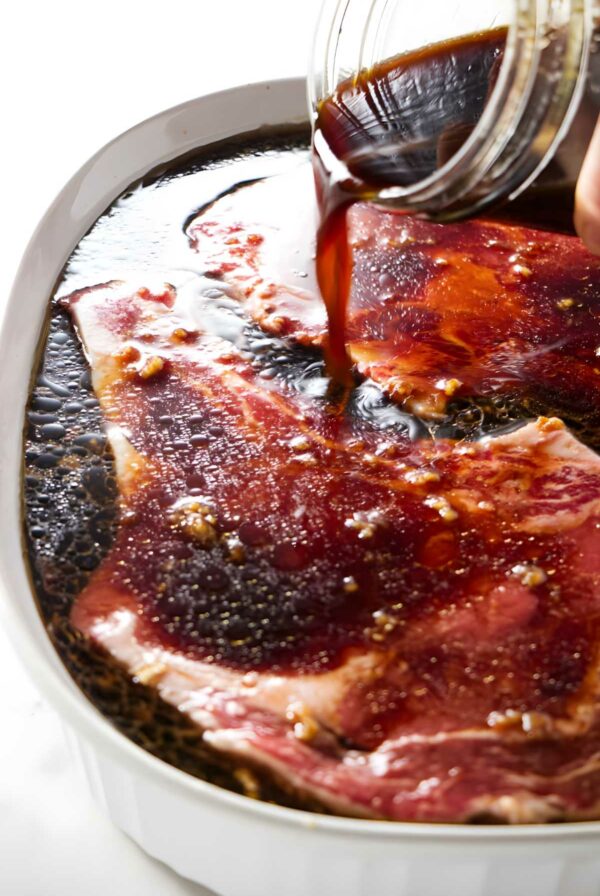 Hand pours dark marinade over raw beef steaks.