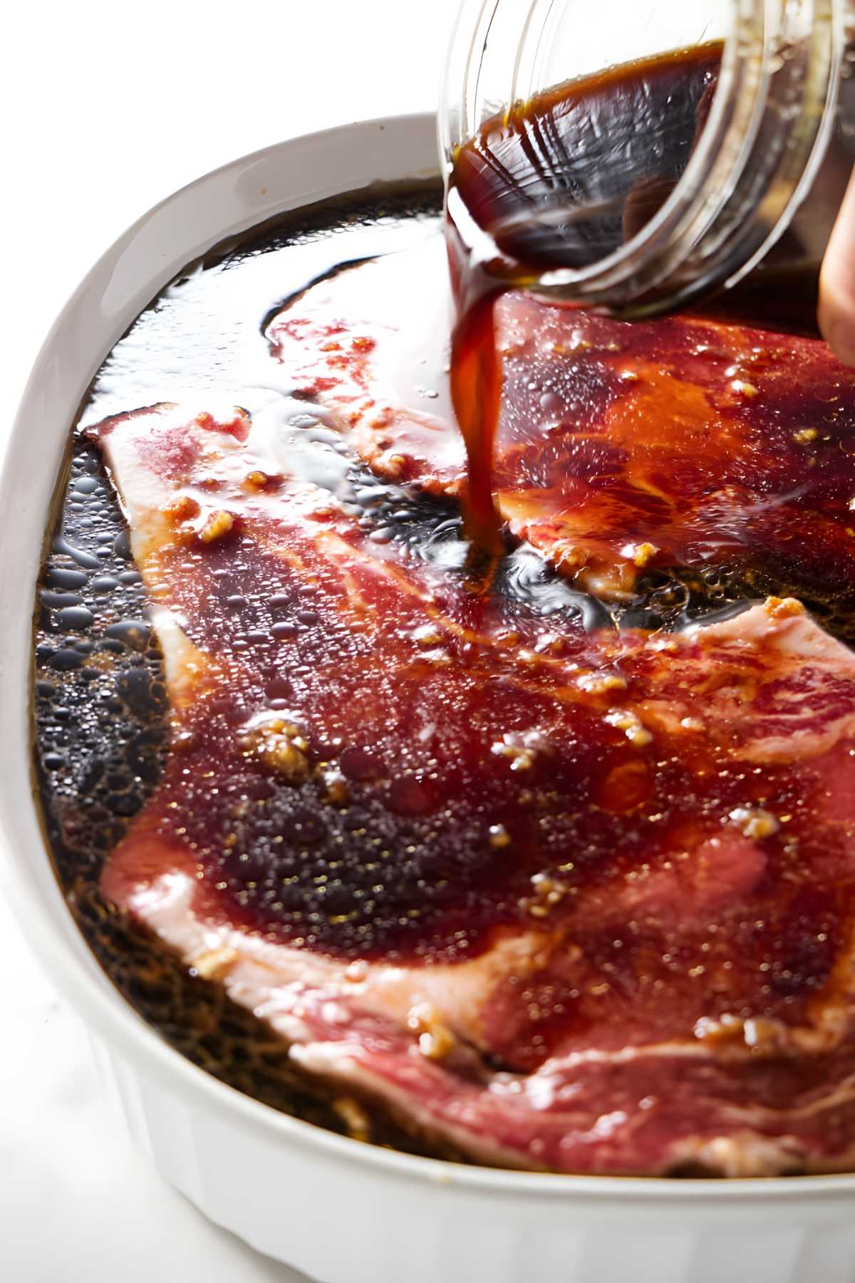 Hand pours dark marinade over raw beef steaks with garlic and spices.