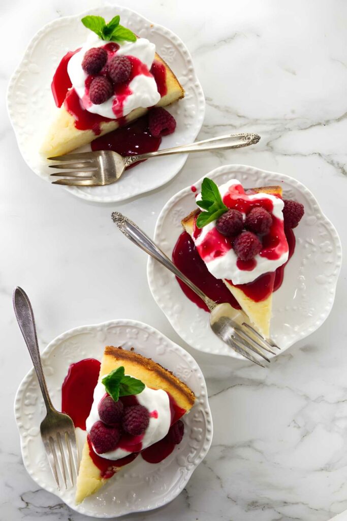 Three cheesecake slices with whipped cream, raspberry sauce, mint, and forks.