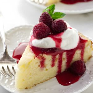 Cheesecake slice with whipped cream, raspberry sauce, raspberries, mint, and fork.