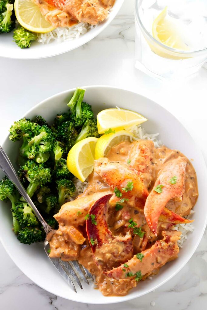 Lobster in tomato sauce with broccoli, lemon wedges, and parsley garnish.