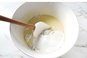 White bowl mixing batter and flour with spatula on marble countertop.