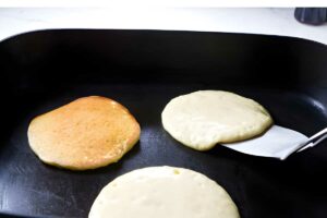 Spatula lifts batter pancake beside cooked and cooking pancakes on griddle.