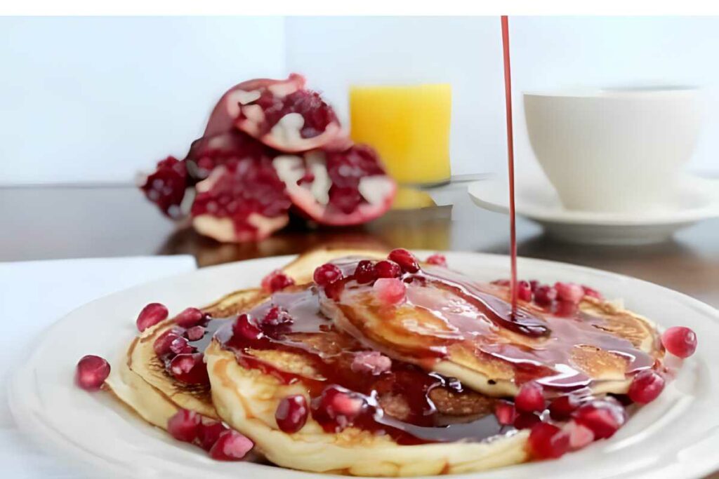 Stack of pancakes with pomegranate seeds and syrup, breakfast drinks nearby.
