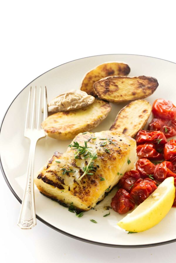 Pan seared sea bass fillet with potatoes, cherry tomatoes, herbs, and lemon wedge.